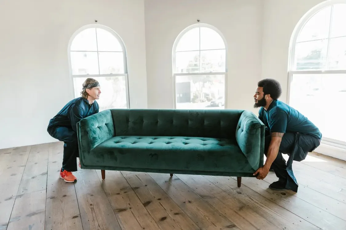 Two movers lifting a green sofa in a bright, emptied room during a home cleanout